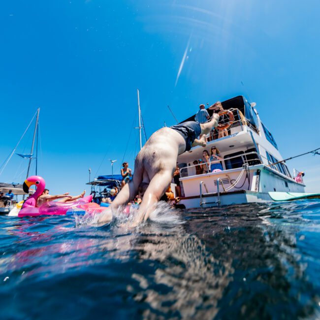 A man dives off a boat into the water. Nearby, people relax on pink inflatable pool floats. Several boats are anchored around, with clear blue skies in the background. The atmosphere is lively and summery.