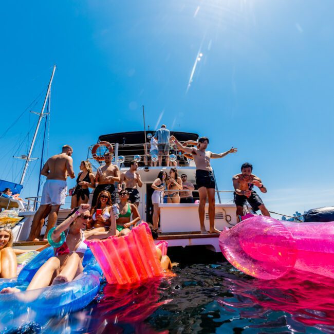 A group of people enjoy a sunny day on a yacht, surrounded by inflatable pool toys, including a large pink flamingo. They are smiling and appear to be having fun in the water. The sky is clear and blue.