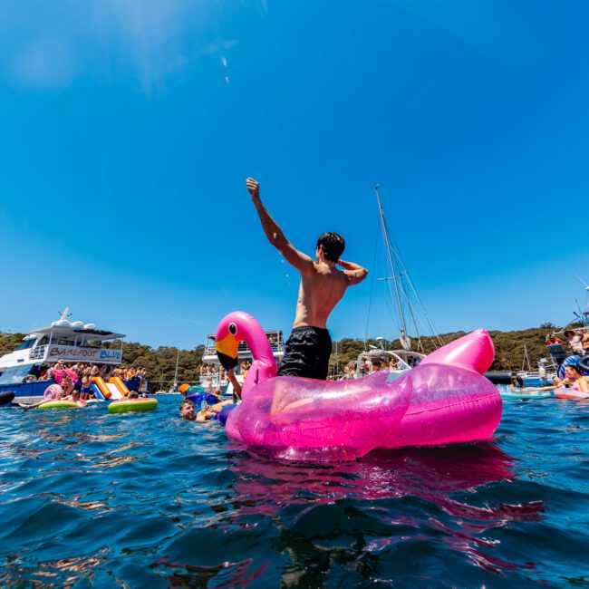A person stands triumphantly on a large, pink flamingo float in a lively water scene surrounded by boats and people enjoying a sunny day. The sky is clear and blue, accentuating the festive atmosphere.