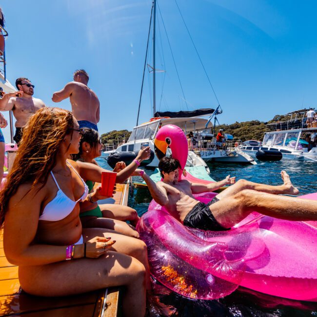 A group of people enjoying a sunny day on a dock and in the water. Some are lounging on pink inflatable floaties, while others are socializing on a yacht. Surrounding them are boats and a clear blue sky.