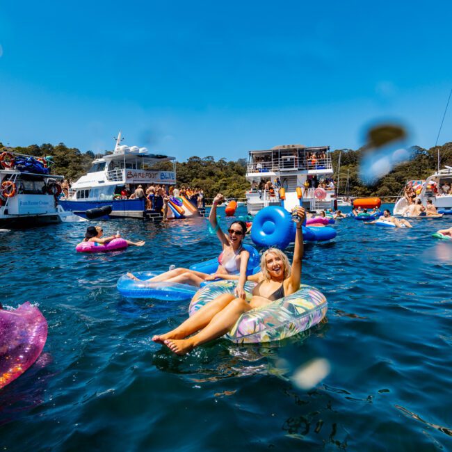 A group of people relax on colorful inflatable floats in a vibrant blue ocean. Boats are anchored nearby, and the sky is clear and sunny. Everyone appears to be enjoying a festive, social atmosphere on the water.
