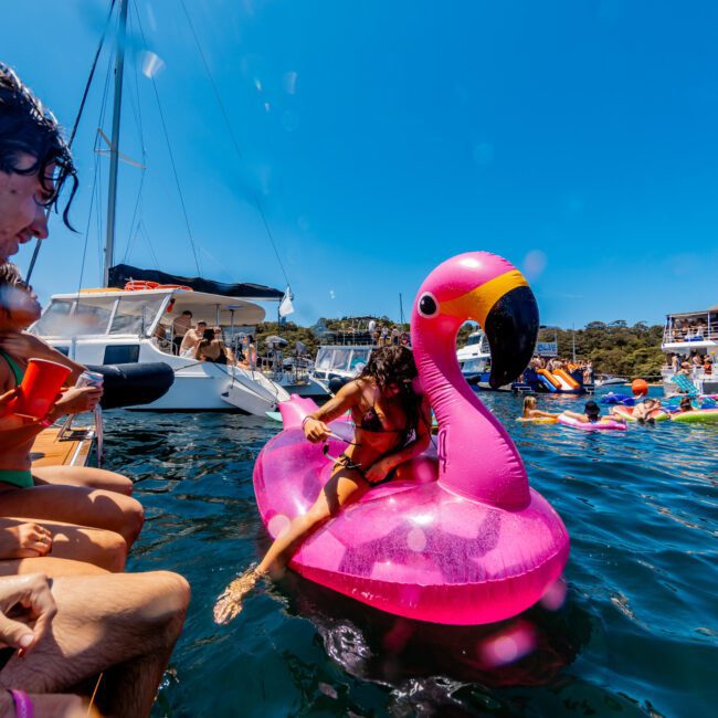 A person sits on a large pink flamingo float in the water, surrounded by boats and other colorful inflatables. A yacht and people enjoying the sun are in the background. Bright, clear sky overhead.