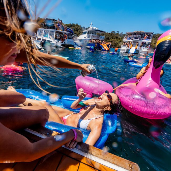 A person in a white bikini lounges on a blue inflatable float in the water, being splashed playfully by someone holding a can. The scene is lively, with colorful inflatables and boats in the background under a bright blue sky.