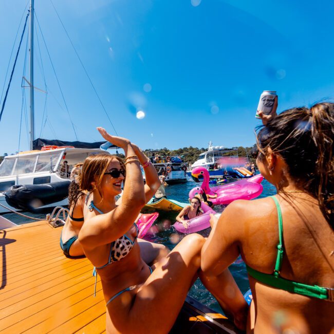 A group of people enjoying a sunny day on the water. Some are on a boat while others float on inflatable toys, including a pink flamingo. The atmosphere is festive and relaxed, with clear blue skies above.