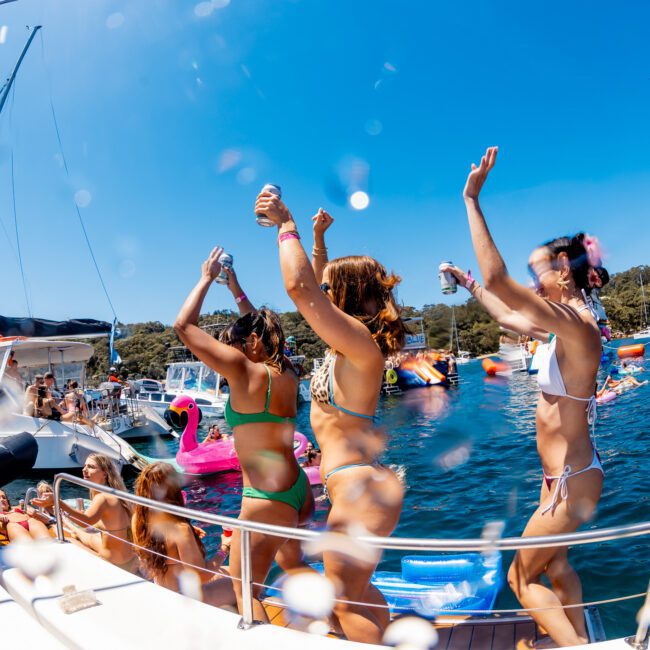 A group of people in swimwear are dancing and holding drinks on a boat under a clear blue sky. Other boats and inflatable toys are visible in the background, floating on a calm body of water.