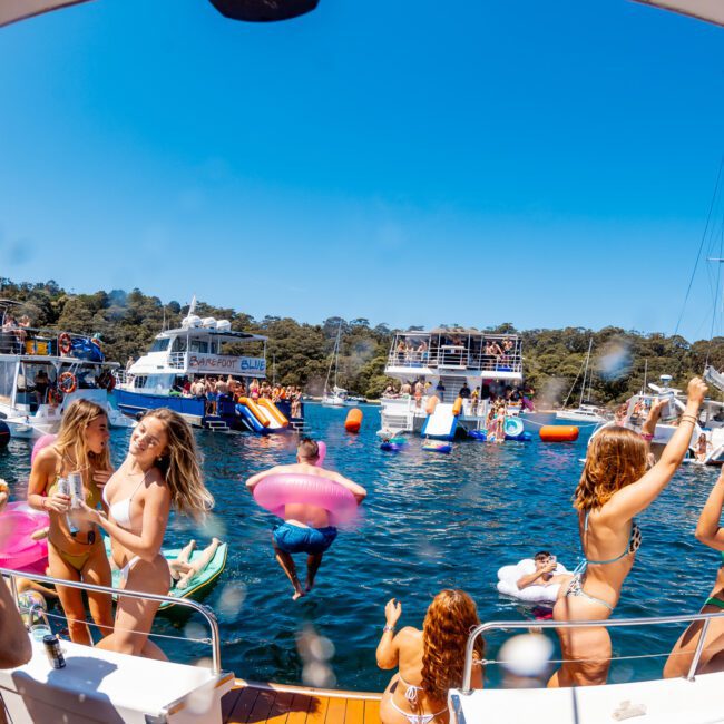 A lively boat party on the water with people dancing and enjoying the sun. Inflatable toys float nearby, and multiple boats can be seen in the background. The scene is vibrant and full of energy with clear blue skies overhead.