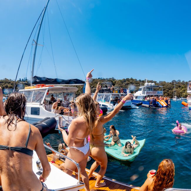 A lively summer scene on a sunny day with people enjoying a boat party. Several individuals are on a boat deck, some dancing, while others relax on inflatable floats in the water. Sailboats and yachts can be seen in the background.