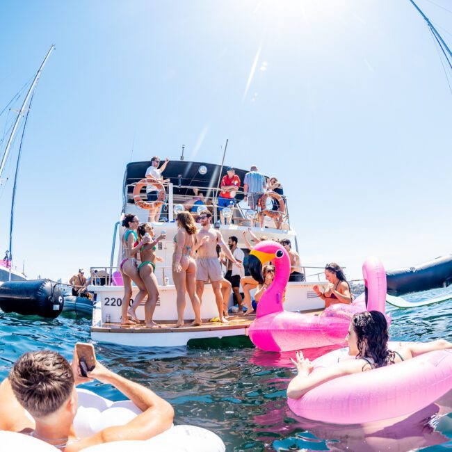 A lively scene at a marina features a group of people in swimsuits enjoying a yacht party. Several individuals are on inflatable floats, including a pink flamingo, in the water surrounded by moored boats on a sunny day.