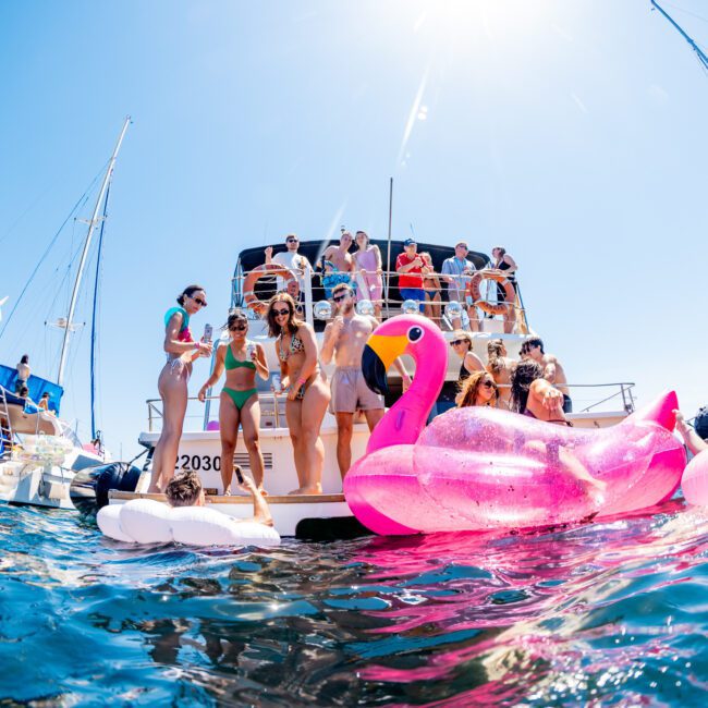 People enjoying a sunny day on a yacht, with some swimming and others on deck. Bright pink flamingo floaties are in the water. Sails and blue sky in the background create a festive atmosphere.