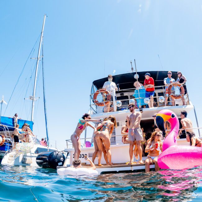 A group of people enjoys a sunny day on a yacht, swimming and lounging with a large inflatable pink flamingo in the water. Other yachts are visible, and the sky is clear and blue.