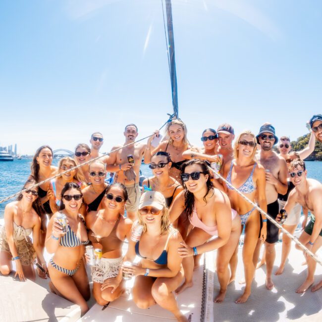 A group of people in swimwear smiling on a boat with a city skyline and blue sky in the background. Some are holding drinks, and everyone is gathered closely on the deck, enjoying a sunny day.
