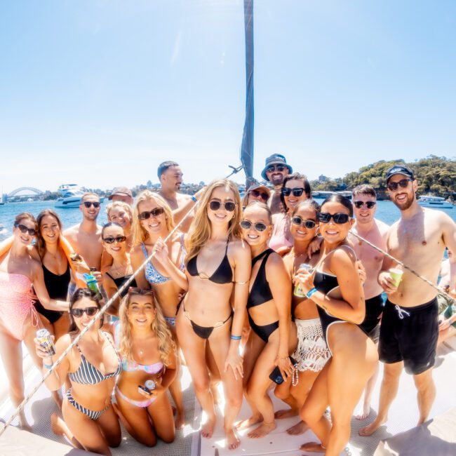 A group of people in swimwear posing together on a boat with a sunny blue sky and city skyline in the background. They are smiling and appear to be enjoying a day out on the water.
