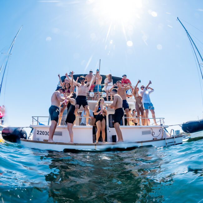 A group of people having fun on a boat under a clear, sunny sky. They are standing, dancing, and socializing on the deck, surrounded by water and other boats nearby. Sunlight reflects on the water, adding a vibrant, summery atmosphere.