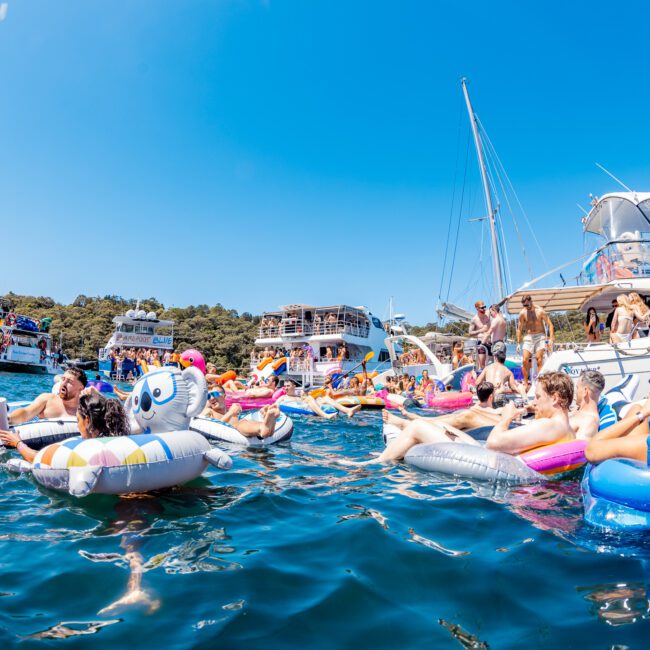 A vibrant summer scene with people relaxing on inflatable floats in a blue, sunny water area. Several boats are anchored nearby, and the sky is clear. The atmosphere is festive and social.