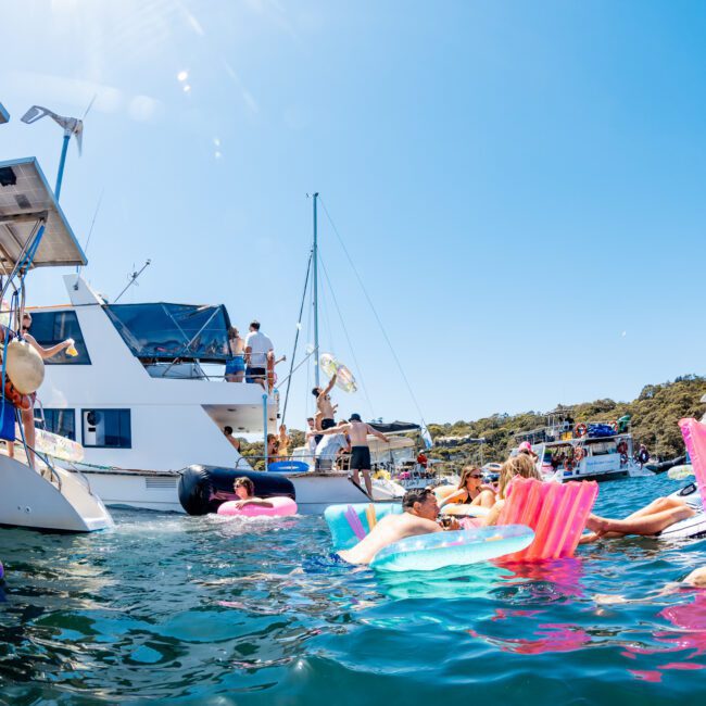 A lively scene of people enjoying a sunny day on the water. Several individuals float on colorful inflatables near a docked boat. Others are gathered on the boat, surrounded by clear blue waters and lush green hills.
