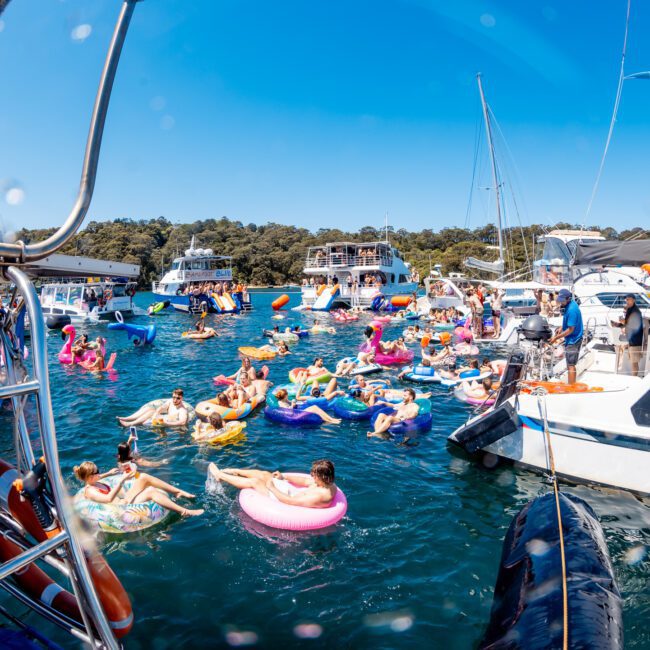 A group of people enjoying a sunny day on a lake, floating on colorful inflatables. Several boats are anchored around them, creating a festive and lively atmosphere. The scene is set against a backdrop of lush greenery and clear blue skies.