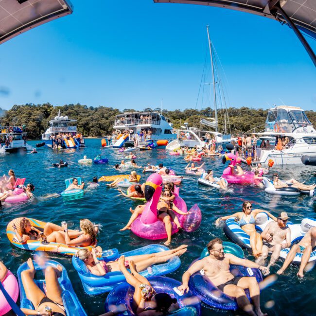 A vibrant scene shows people enjoying a sunny day on the water. They are lounging on various colorful inflatables like flamingos and floats. Several boats are anchored nearby, and the sky is clear and blue.