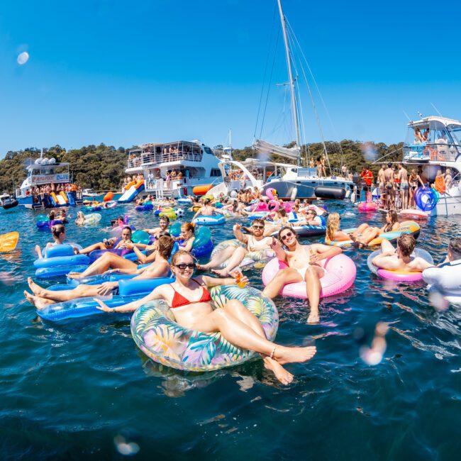 A group of people relax on colorful inflatable floats in a sunny bay. They are surrounded by boats and enjoying a festive atmosphere in the water. The scene is lively and vibrant, with clear blue water and bright skies.