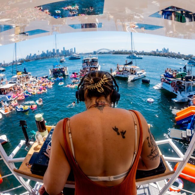 Person with headphones DJs on a boat overlooking a bustling waterfront scene. Multiple boats and colorful floats fill the water, with a view of the city skyline in the background.