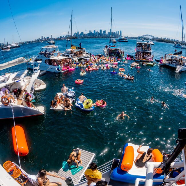 A lively scene on a sunny day with numerous boats gathered in the water. People are enjoying themselves on inflatable floats and swimming. In the background, a city skyline is visible under a clear blue sky.