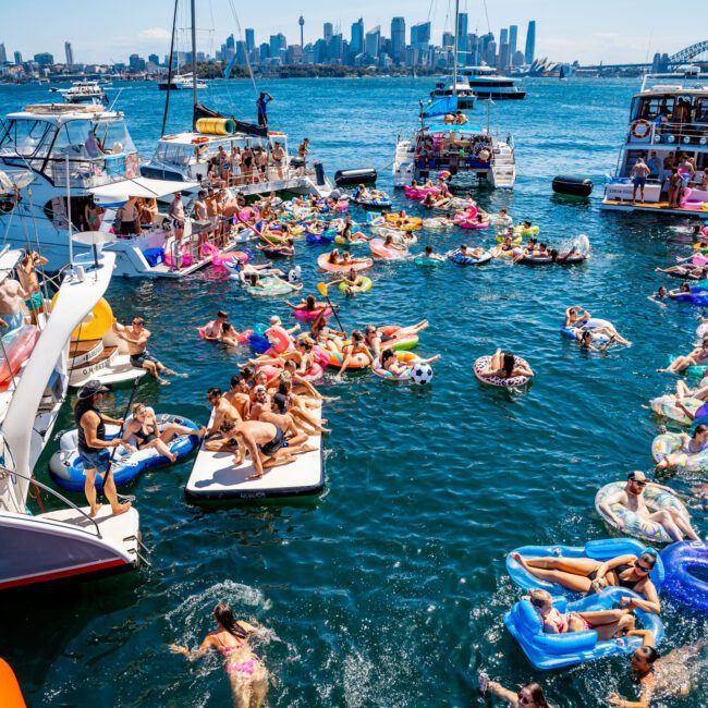 A lively scene of people enjoying a sunny day on a harbor. Many are swimming, floating on colorful inflatables, and relaxing on boats. The city skyline is visible in the background under a clear blue sky.