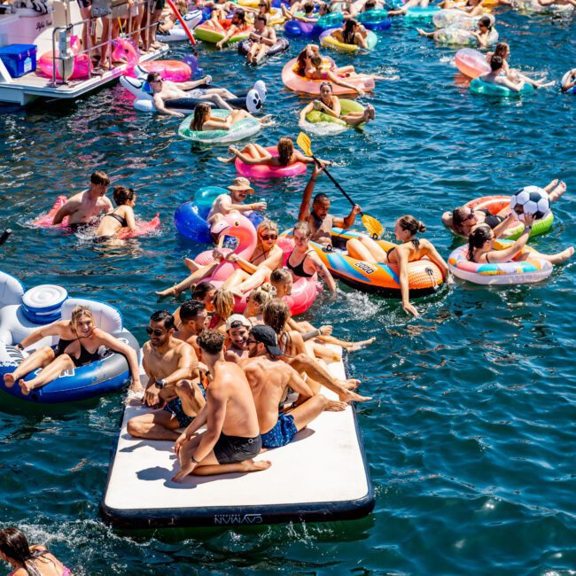 A large group of people enjoying a sunny day on a lake, relaxing on inflatable floats and a platform. Boats are lined up in the background. The scene is lively, with colorful inflatables and swimmers in the water.