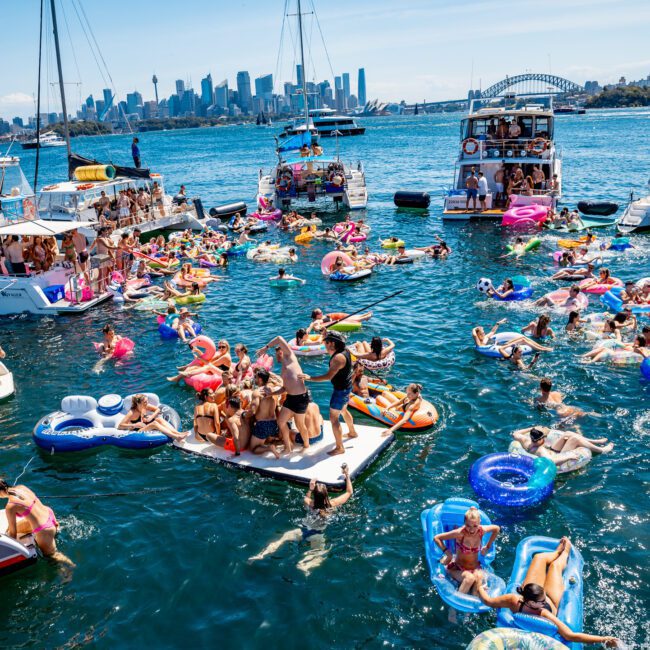 A lively scene on the water with people enjoying a sunny day on inflatable floats, surrounded by boats. The city skyline is visible in the background.