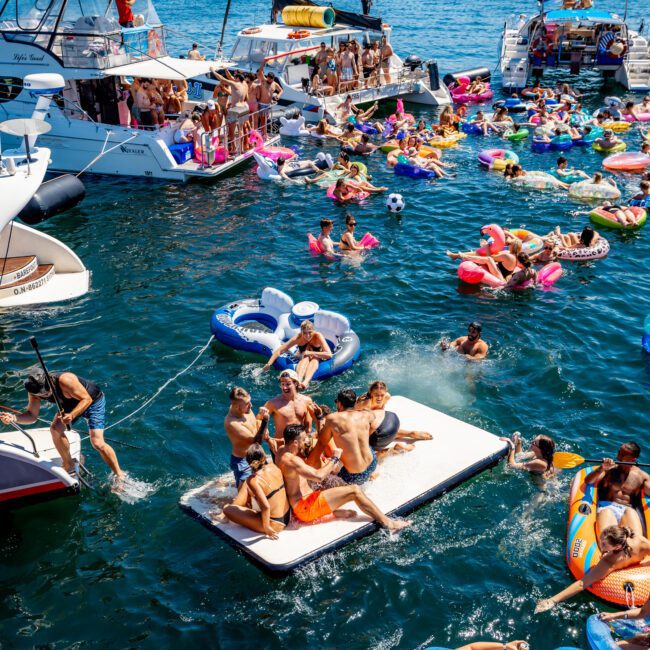 People enjoying a sunny day on a harbor, surrounded by boats and floating inflatables. The background features a city skyline. The scene is lively with individuals swimming, socializing, and relaxing on the water.