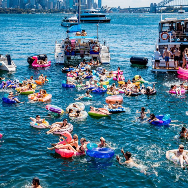 People enjoying a sunny day floating on colorful inflatables in a bay, with boats anchored nearby. The city skyline and a distinctive bridge are visible in the background.