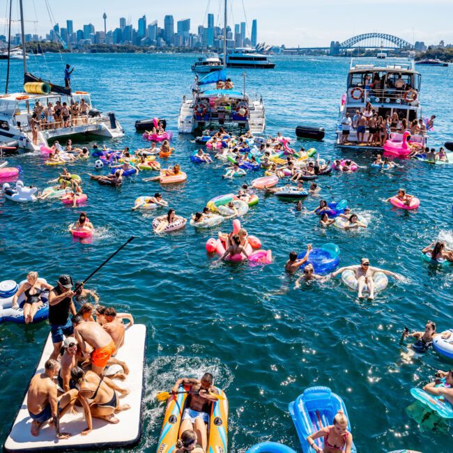 A crowded party scene on blue water with people on colorful inflatables, surrounded by anchored boats. The skyline and an arch bridge are visible in the background on a sunny day.