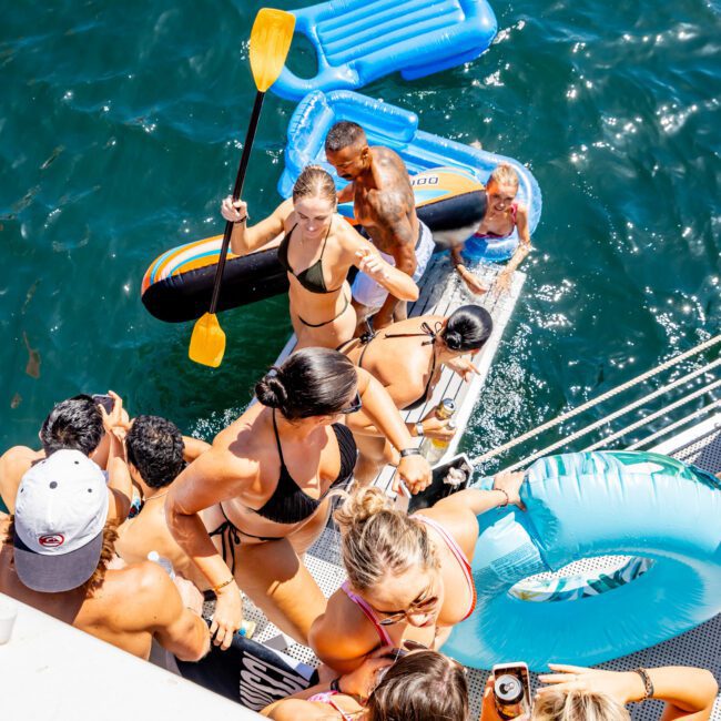 A group of people enjoy a sunny day on a boat and in the water. Some are on a float with paddles, while others relax in swimsuits. An inflatable lounge and ring float nearby. The water is a deep blue, and the scene is lively and festive.