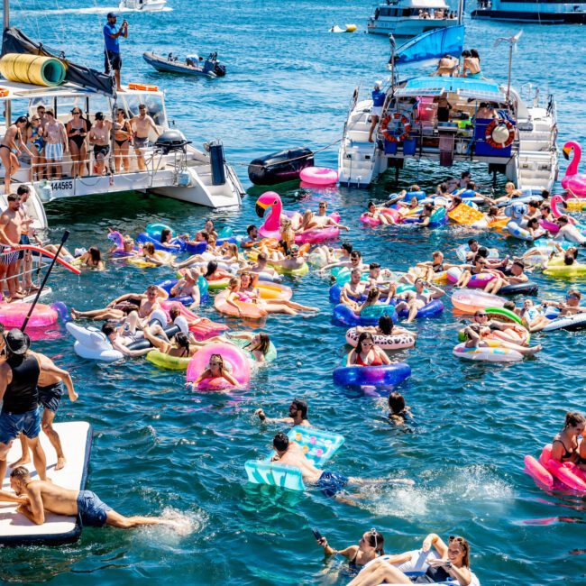 A lively scene of people enjoying a sunny day on colorful inflatables in the water, with boats nearby. The city skyline is visible in the background.