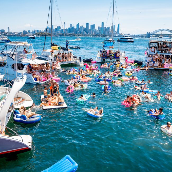 Boats are anchored in a harbor with people on inflatable rafts and pool floats enjoying a party in the water. The city skyline is visible in the background.
