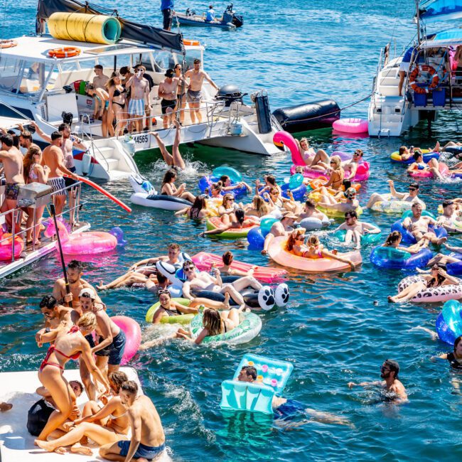 A lively scene of people on inflatable floats in the water, surrounded by boats. Many are sunbathing and socializing under a bright blue sky. The atmosphere is festive and crowded with colorful inflatables and swimwear.