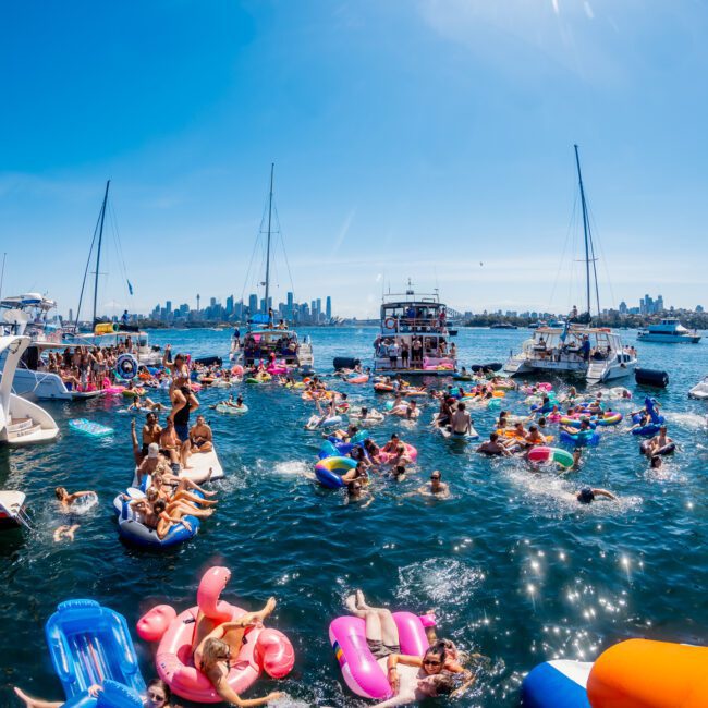 A lively scene of a boat party with people on colorful inflatable floats and boats in a sunny harbor. The city skyline is in the background under a clear blue sky.