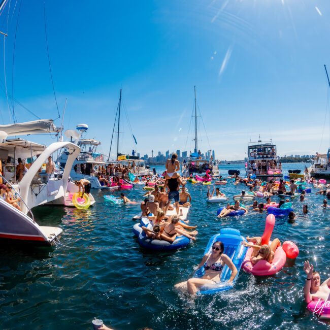 A lively scene on the water with people enjoying a sunny day. Numerous boats and colorful inflatables fill the blue waters, while a city skyline stands in the background. Sunlight sparkles on the water, creating a festive atmosphere.