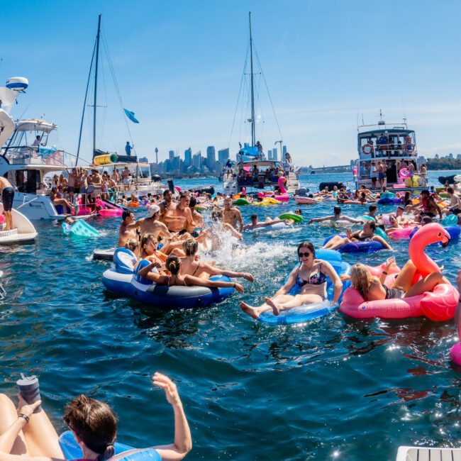 A lively scene on the water with people on colorful inflatables and boats. The sunny sky brightens the gathering, as the city skyline is visible in the background. Everyone is enjoying the warm day and the cool water.