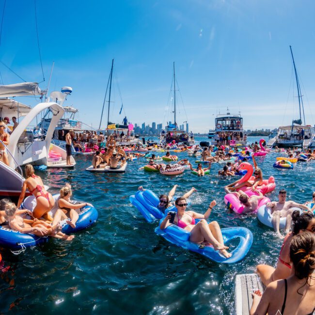 A lively scene of people enjoying a sunny day on the water, floating on inflatable loungers and pool toys near boats. Skyscrapers are visible in the distant background under a clear blue sky.