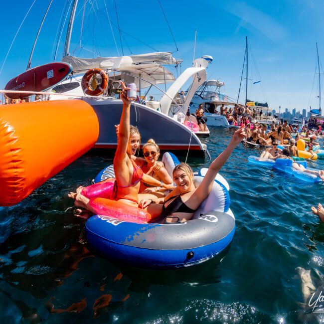 A group of people enjoy a sunny day on the water, floating on inflatables near boats. The scene is lively, with many individuals relaxing and having fun, surrounded by clear blue waters and a city skyline in the background.