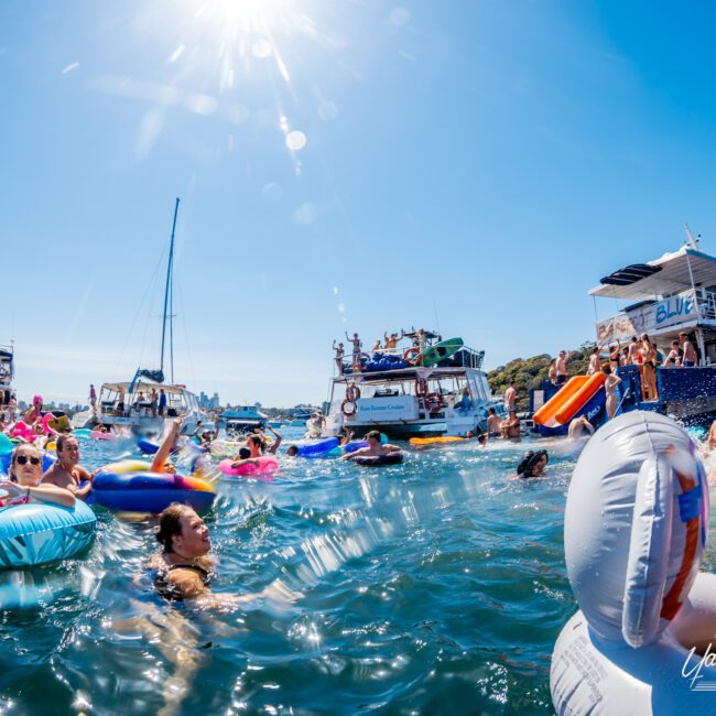 A lively scene of people enjoying a sunny day on the water. Many are swimming or floating on colorful inflatables near several boats. Bright sunlight reflects off the water, adding to the festive atmosphere.