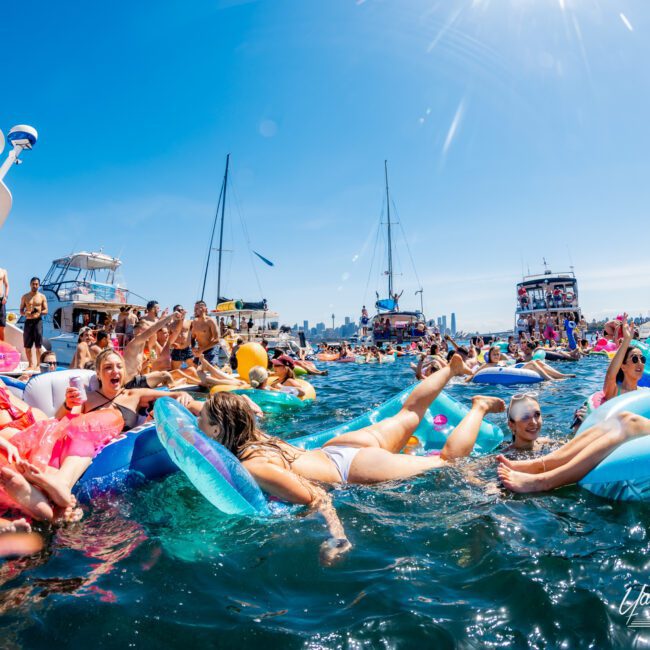 A vibrant scene with people on colorful inflatable rafts enjoying the water near anchored boats. The sun is bright, the sky is clear, and more people are visible on the boats in the background. Everyone is relaxed and having fun.