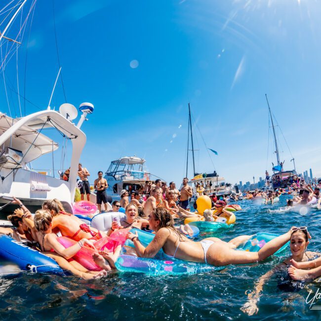 A lively scene at a yacht party with people enjoying themselves in the water on colorful inflatables. Several yachts are anchored nearby under a clear blue sky, and there is a large crowd celebrating and socializing in the background.