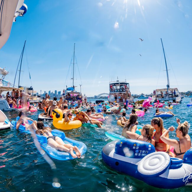 A lively group of people enjoy a sunny day on the water. They're floating on colorful inflatables near boats, with a city skyline and the harbor bridge in the background. The scene is bright and festive.