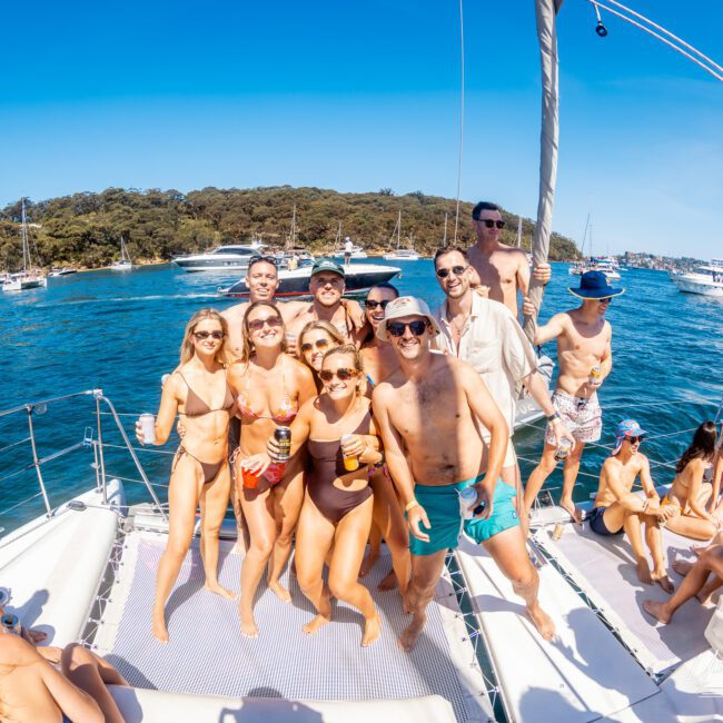 A group of people enjoying a sunny day on a sailboat. They are wearing swimsuits and sunglasses, smiling and posing for the camera. The ocean and other boats are visible in the background under a clear blue sky.