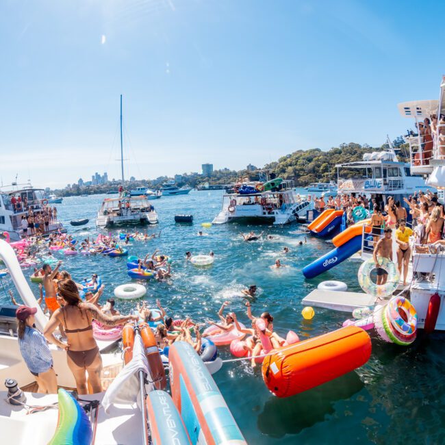 A lively scene on a sunny day with people in swimsuits enjoying a yacht party. Boats are docked close together, and attendees are swimming and using inflatables in the water, creating a festive and vibrant atmosphere.