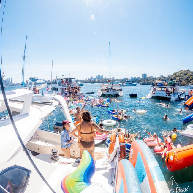 A lively scene of people enjoying a sunny day on boats and colorful inflatables in a bay. The blue sky and distant cityscape frame the background, with music and water activities creating a festive atmosphere.