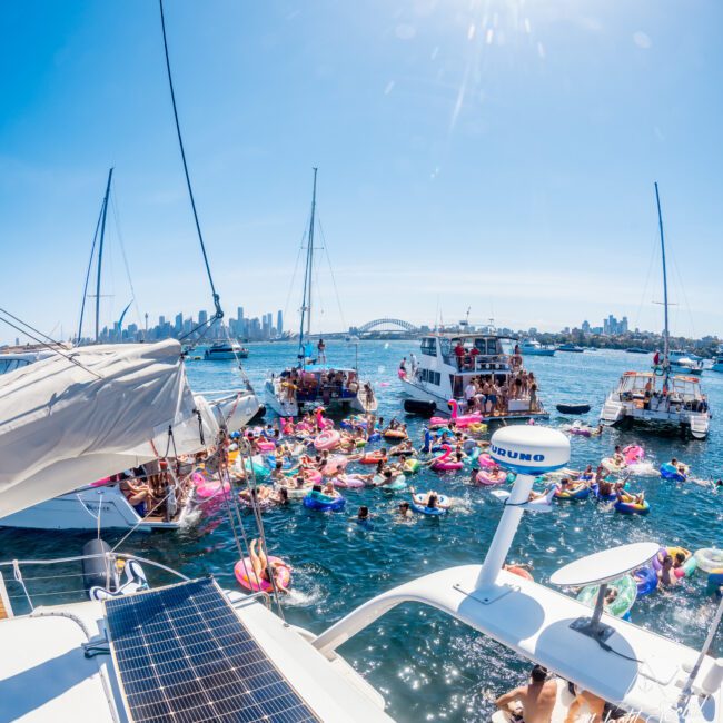 A lively scene of people on inflatable rings enjoying a sunny day on the water surrounded by boats. The skyline and a bridge are visible in the background. The sun shines brightly, enhancing the festive atmosphere.