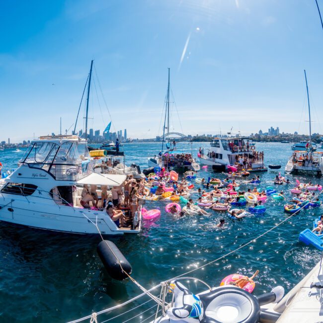 A vibrant scene of a boat party on a sunny day, with numerous people on inflatable floats in the sea, surrounded by several boats. The background features a city skyline with tall buildings under a clear blue sky.