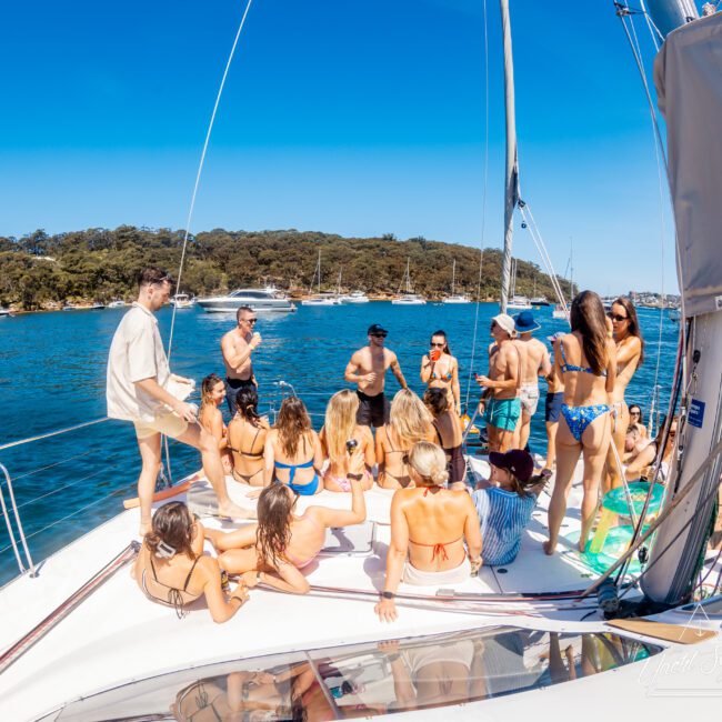A group of people enjoying a day on a sailboat under a clear blue sky. They are wearing swimwear and sitting or standing on the deck, surrounded by calm blue waters and green wooded shores in the background.