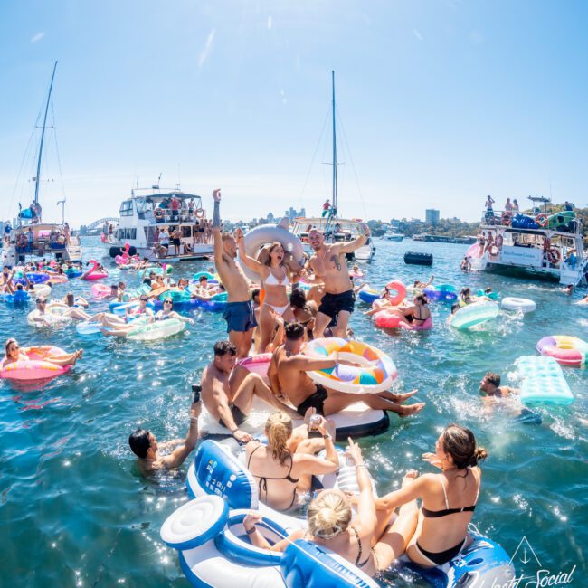 A lively gathering of people enjoying a sunny day on the water. They are on colorful inflatables, with yachts and sailboats in the background. The mood is festive, with clear blue skies and sunlight reflecting on the water.